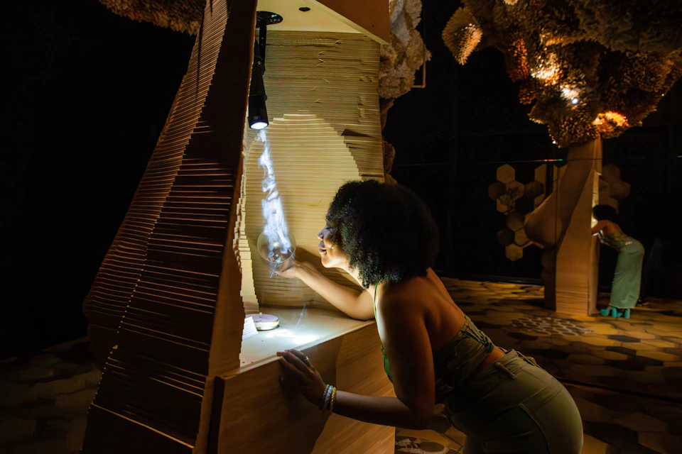 A woman leans into an art installation, inspecting illuminated smoke under a spotlight, surrounded by intricate wooden structures.