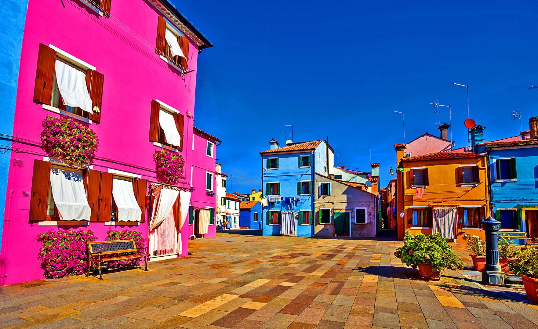 Vibrant street scene with brightly colored houses in pink, blue, yellow, and orange. Curtains and potted plants adorn the windows.