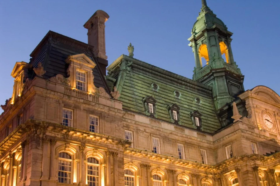 A large, ornate, historic building with illuminated windows and a green-roofed tower against a dusky sky.