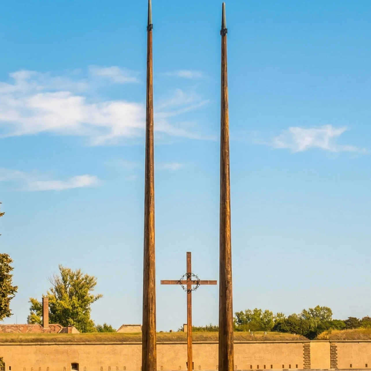 A cross and two tall spires against a blue sky, with trees and a low wall in the background.
