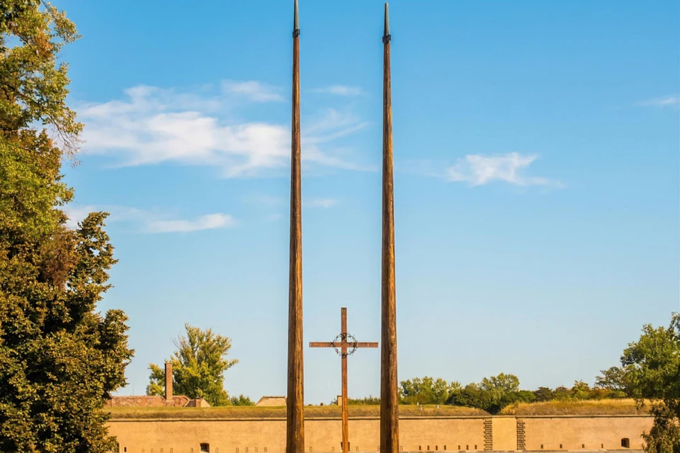 A cross and two tall spires against a blue sky, with trees and a low wall in the background.