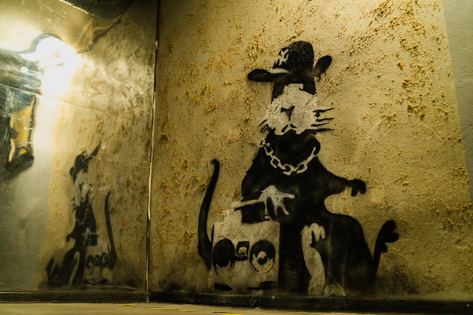 Graffiti of a cat wearing a hat and chain, holding a boombox, is painted on a textured, beige wall.