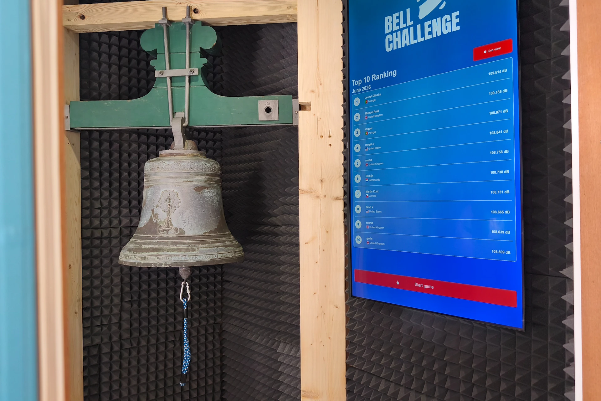 A metallic bell mounted on a wooden frame next to a digital screen displaying a leaderboard for a 'Bell Challenge.'