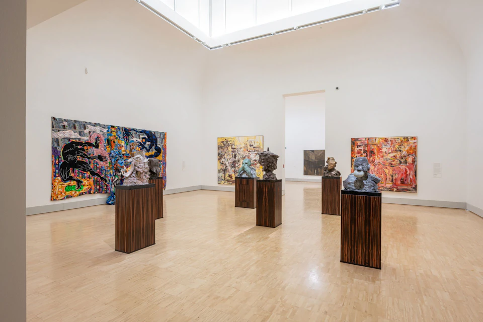 A gallery room with sculptures on wooden pedestals and colorful abstract paintings on the walls.