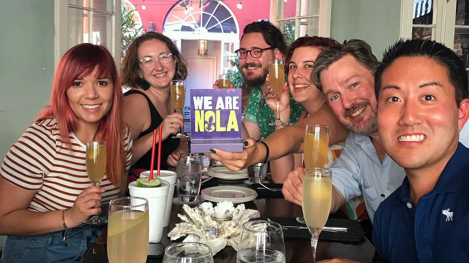 A group of people at a table holding drinks, with one holding a sign reading "We Are NOLA," in a festive setting.