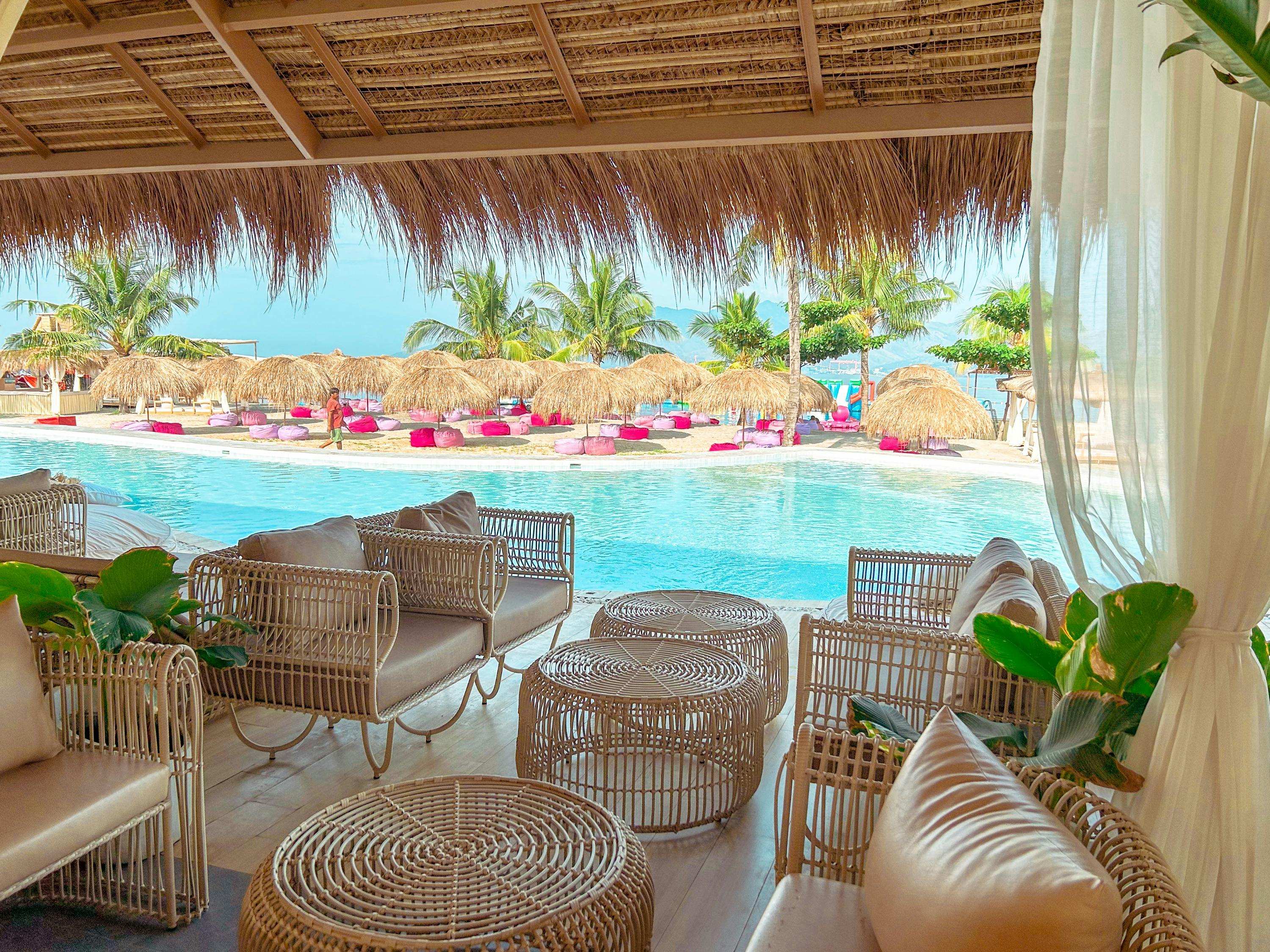 A tropical beachside lounge with wicker furniture, a pool, and a view of thatched umbrellas and palm trees.