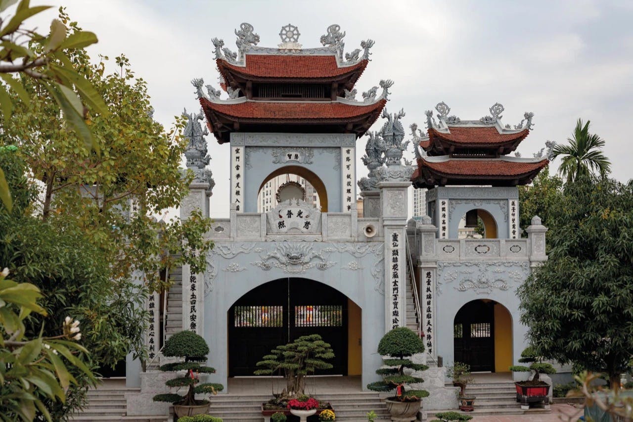Entrée d'un temple asiatique traditionnel avec des pierres ornées, des toits de tuiles rouges et des bonsaïs au premier plan.