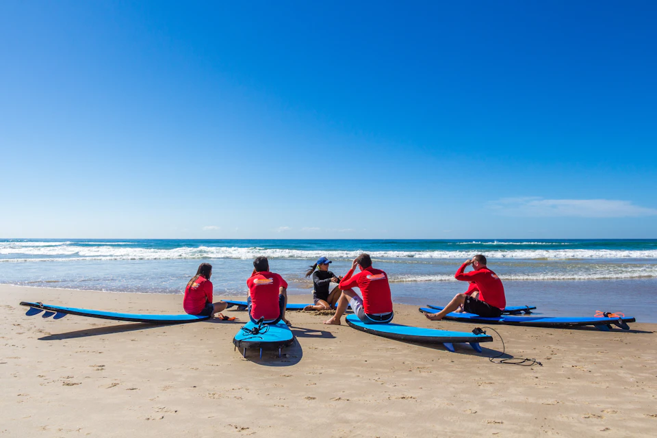 Five people in red shirts sitting on a beach with surfboards, facing the ocean under a clear blue sky.