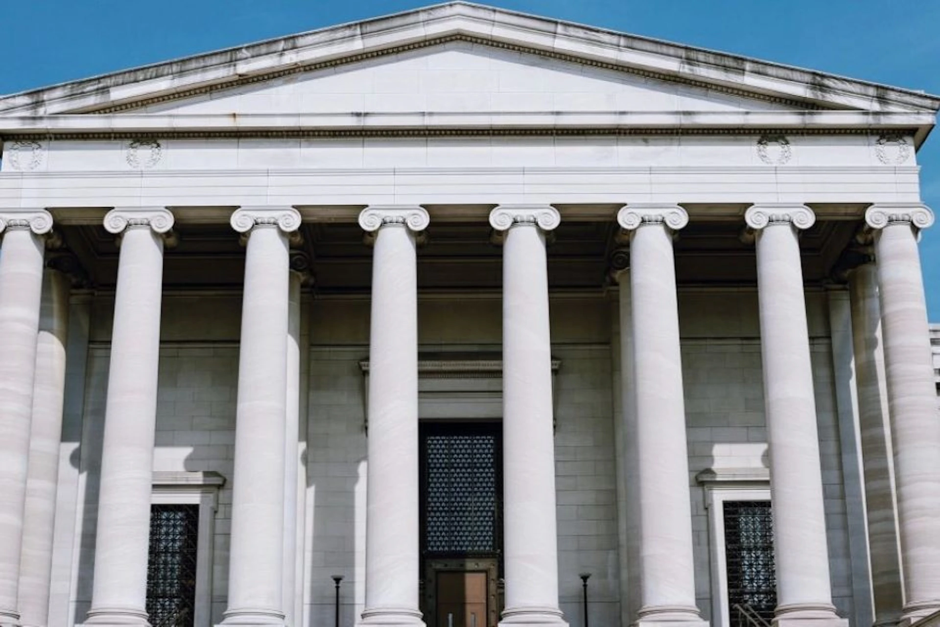 A neoclassical building facade with six large columns supporting a pediment, featuring a main entrance with ornate grilles on the doors.
