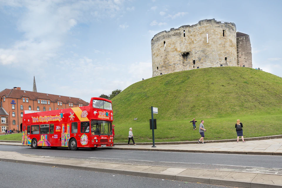 A red double-decker tour bus drives past an old stone tower on a grassy hill with pedestrians walking nearby.