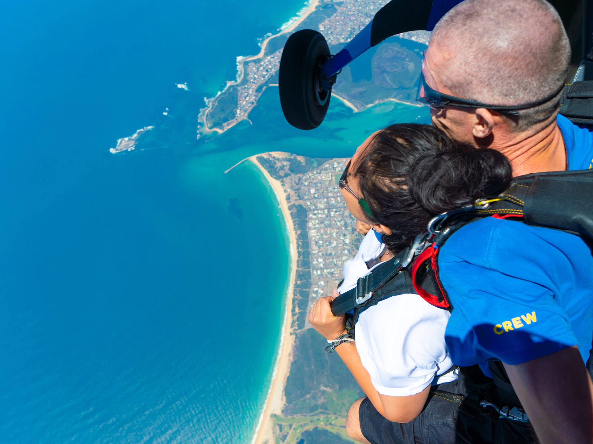 Two people tandem skydiving above a coastal town and beach, with blue ocean and coastline visible below.