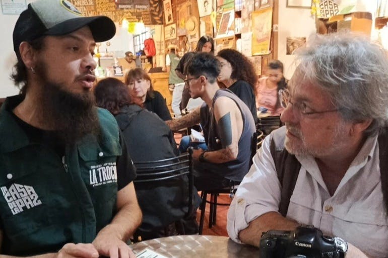 Živá kavárenská scéna s lidmi, kteří mluví a komunikují u různých stolů. V centru pozornosti jsou dva muži, zabývající se konverzací.