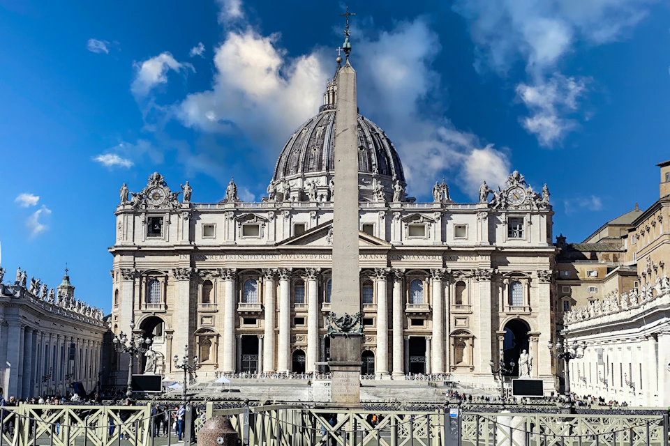 A view of St. Peter's Basilica with its dome and an obelisk in the foreground under a partly cloudy blue sky.