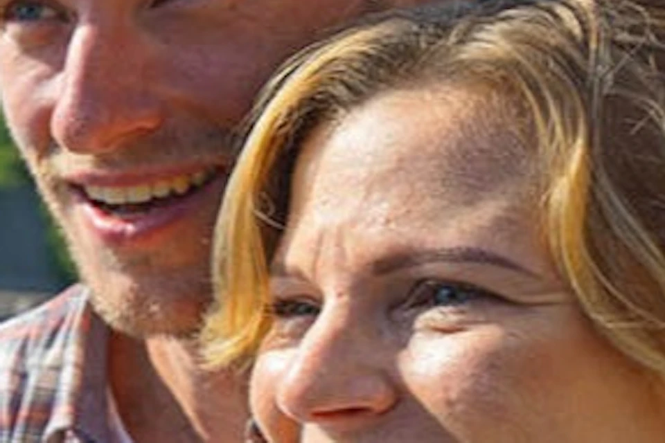 A smiling couple on a railing overlooking a picturesque lakeside village with mountains and a church steeple in the background.