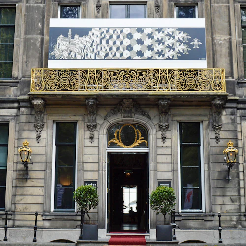 Entré till en historisk byggnad med en gyllene utsmyckad balkong, flankerad av två lampor, träd i krukor och en röd matta som leder inuti.