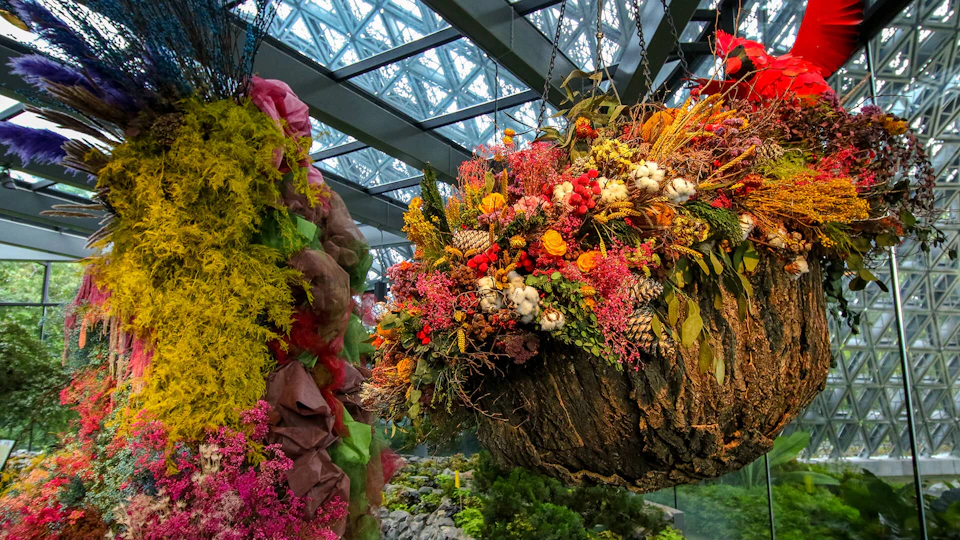 Hanging floral arrangement with brightly colored flowers and foliage in a greenhouse with a glass roof.