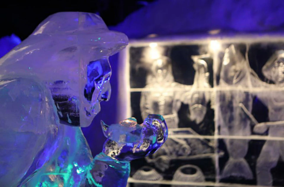 A close-up of an ice sculpture of a person holding an object, with etched artwork on ice in the background. Blue and purple lighting.
