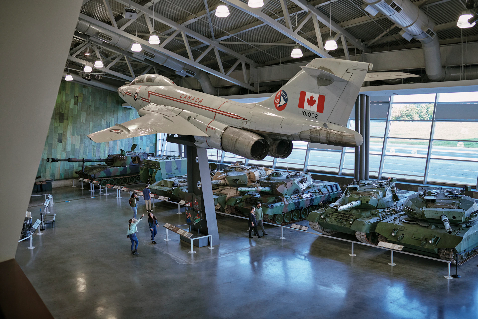 Indoor museum with a Canadian fighter jet on display above military tanks. Visitors walk around exhibits.