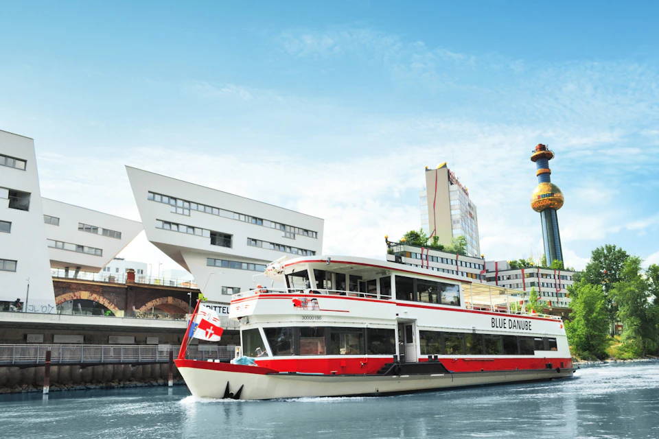 A white and red boat named "Blue Danube" with Swiss flag on a river, with modern buildings and a blue sky in the background.