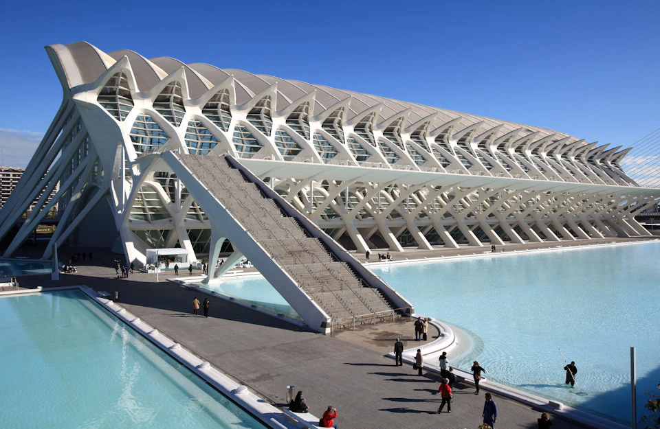 Modern white building with arched structures, large staircase, and people walking near reflective pools under a clear blue sky.