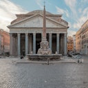 Edificio storico con colonne, una fontana con obelisco al centro, circondato da una pavimentazione in ciottoli e affiancato da edifici antichi.