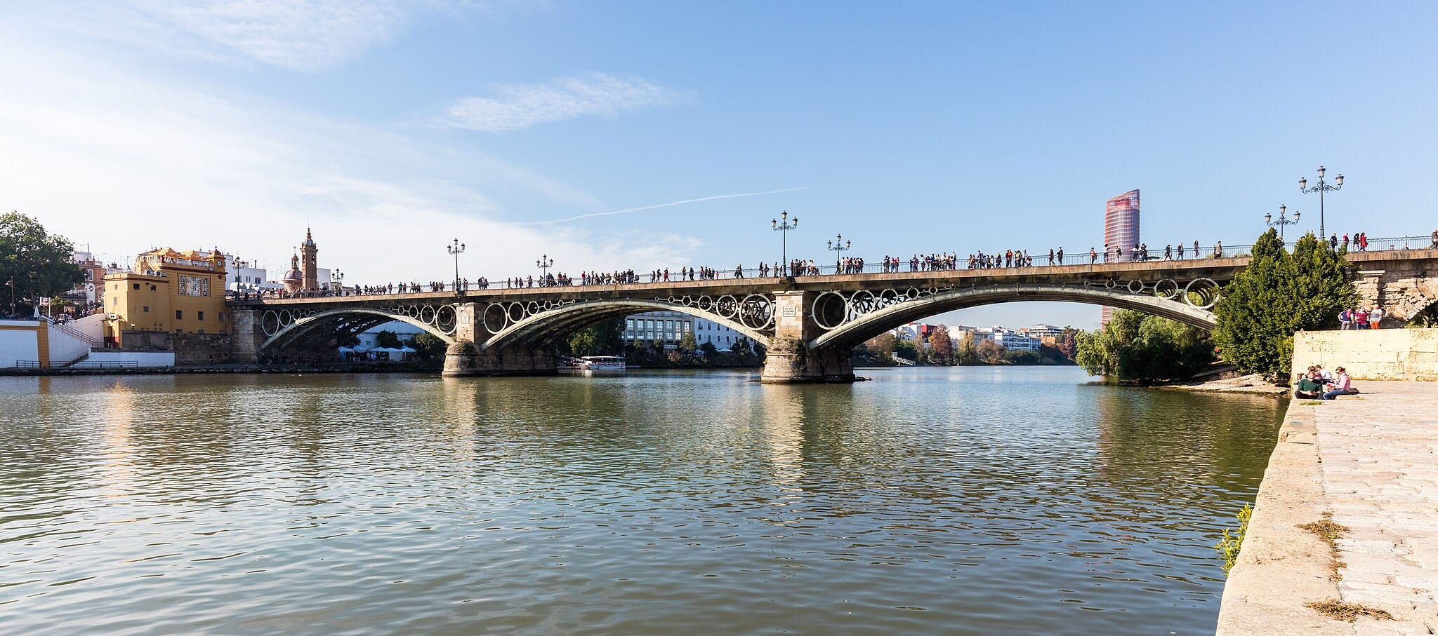 Un gran pont ornamentat travessa un riu amb gent caminant per ell. Els edificis i els arbres són visibles al fons.