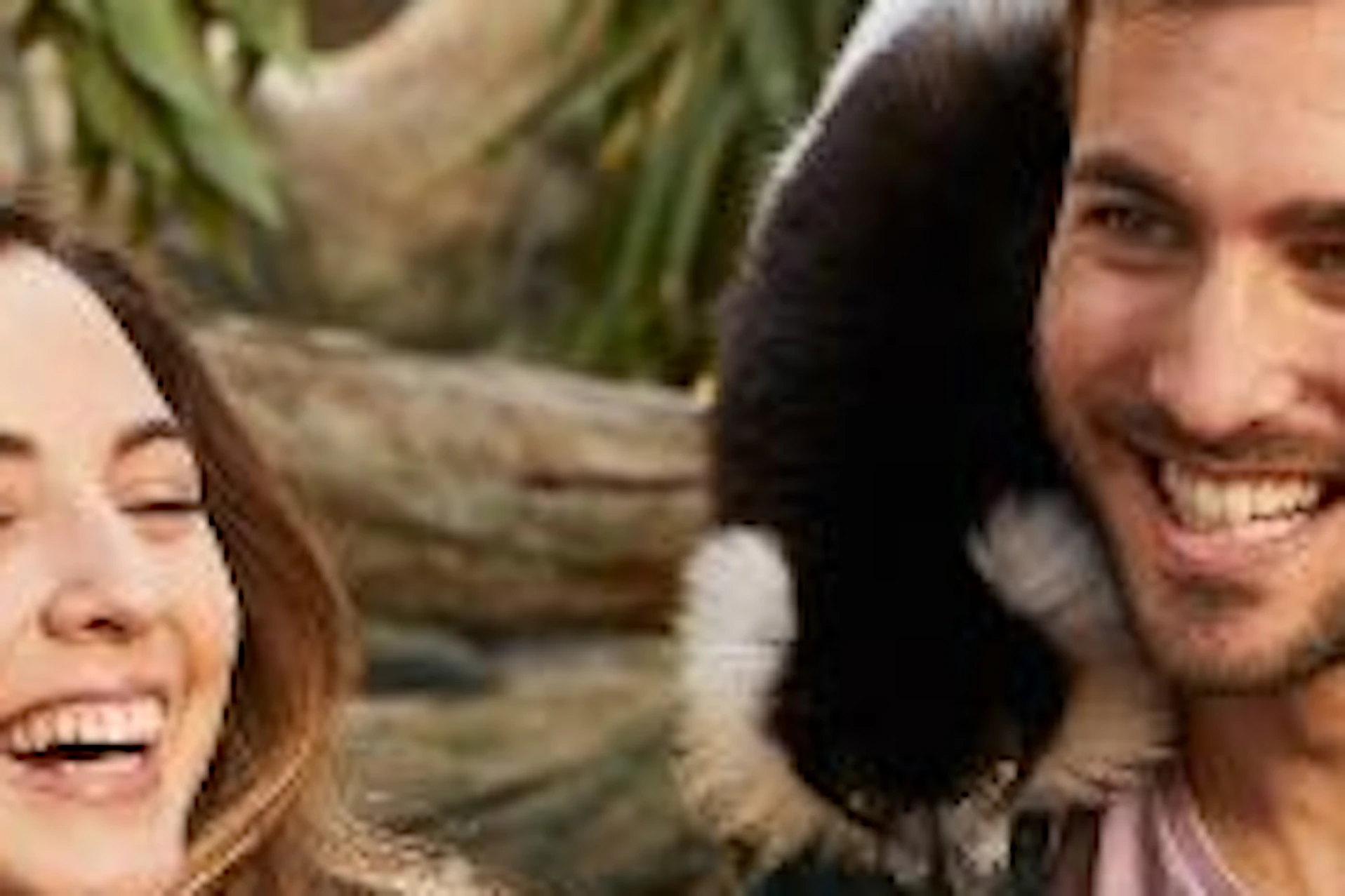 A man and a woman are smiling whilst holding black and white lemurs, with foliage and tree branches in the background.