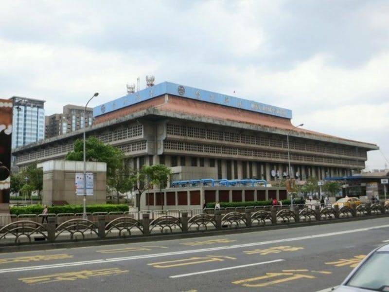 Ένα μεγάλο, πολυώροφο κτίριο με κόκκινη στέγη, ορατά παράθυρα και μια πινακίδα σε ξένη γλώσσα, που βρίσκεται κατά μήκος ενός δρόμου με σημάνσεις.