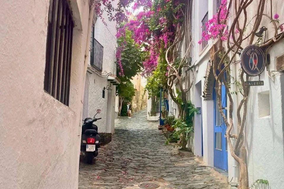 A narrow, cobbled street lined with white buildings, blooming pink flowers and a parked scooter.