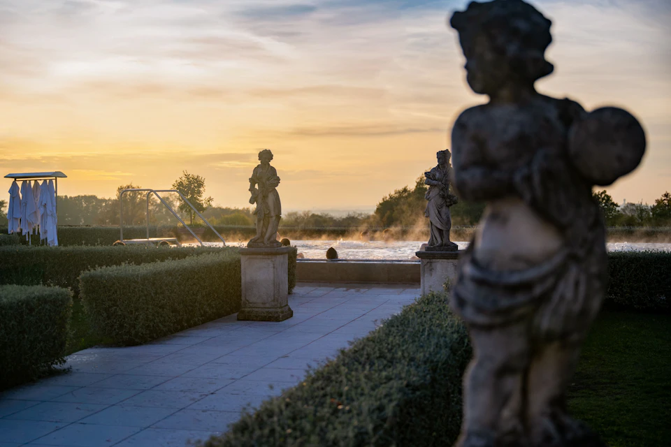 A serene outdoor scene with stone statues, trimmed hedges, and a pool at sunrise, creating a tranquil atmosphere.