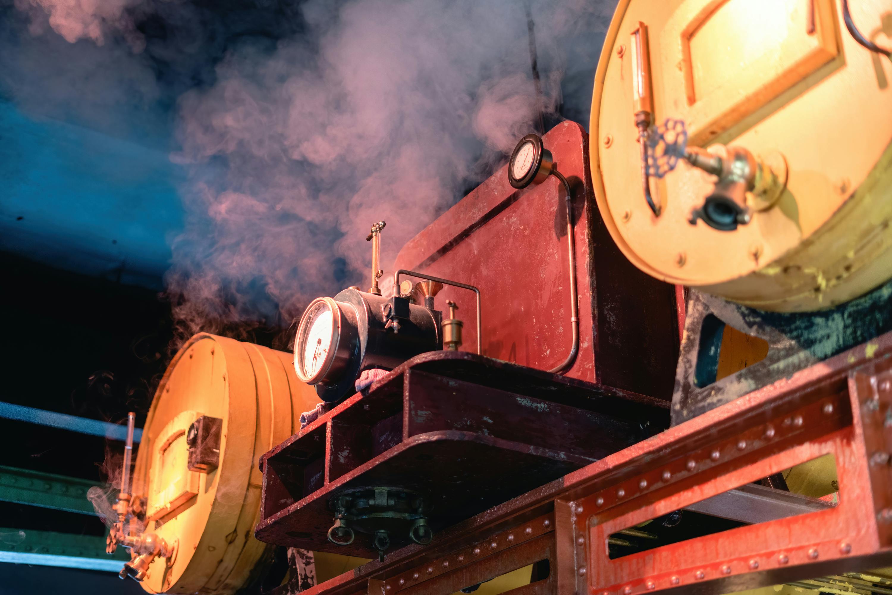 조명이 희미한 환경에서 게이지, 파이프 및 대형 원통형 부품을 특징으로 하는 증기를 방출하는 산업용 기계.