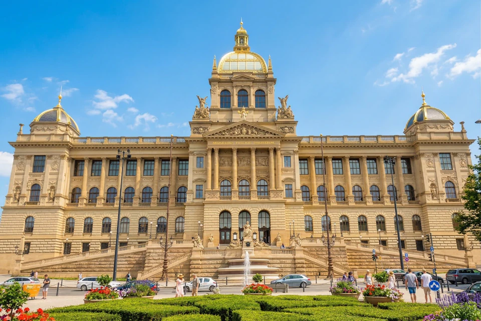 A grand historic building with a golden dome, statues, and stairs, set against a clear blue sky, with people and cars nearby.