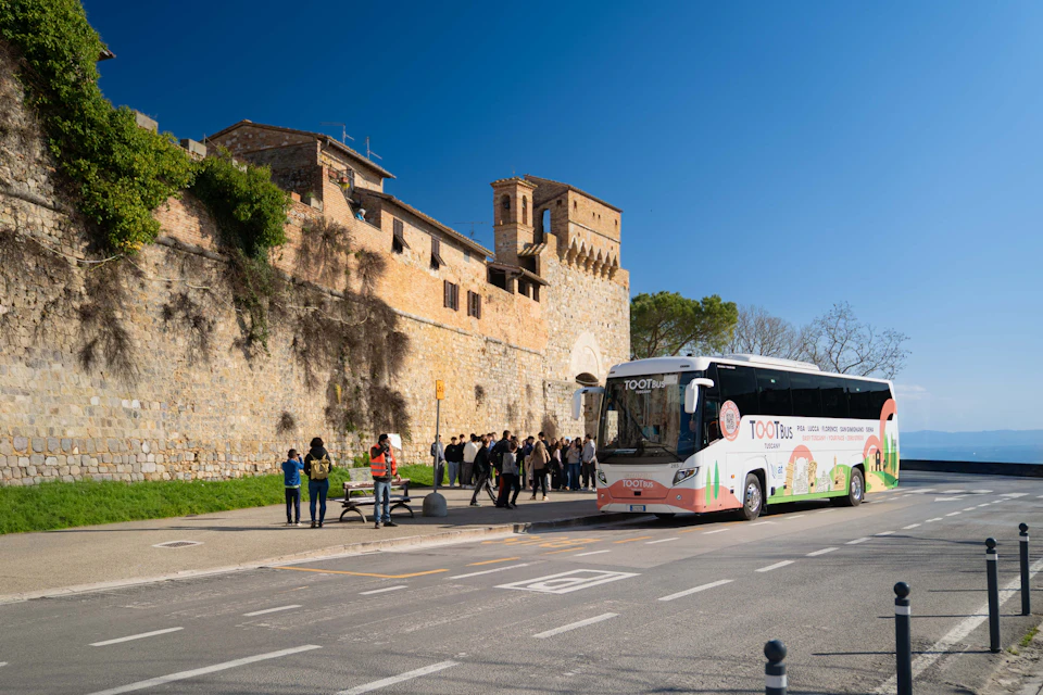 A tour bus parked near a historic stone building under a clear blue sky, with people walking and gathering nearby.