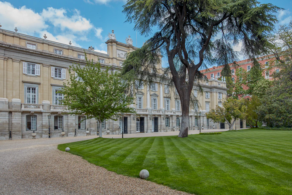 Manicured garden with gravel paths, large trees, and well-maintained grass in front of a historic, multi-story building.