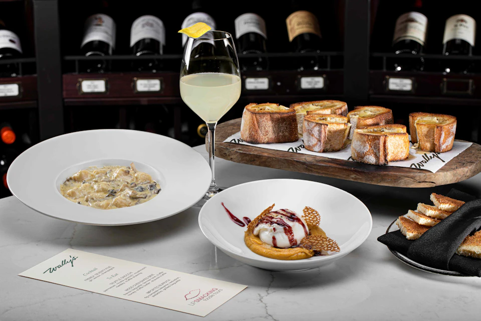 A table with a plate of pasta, a dessert, garlic bread, a lemon cocktail, a menu, and wine bottles in the background.