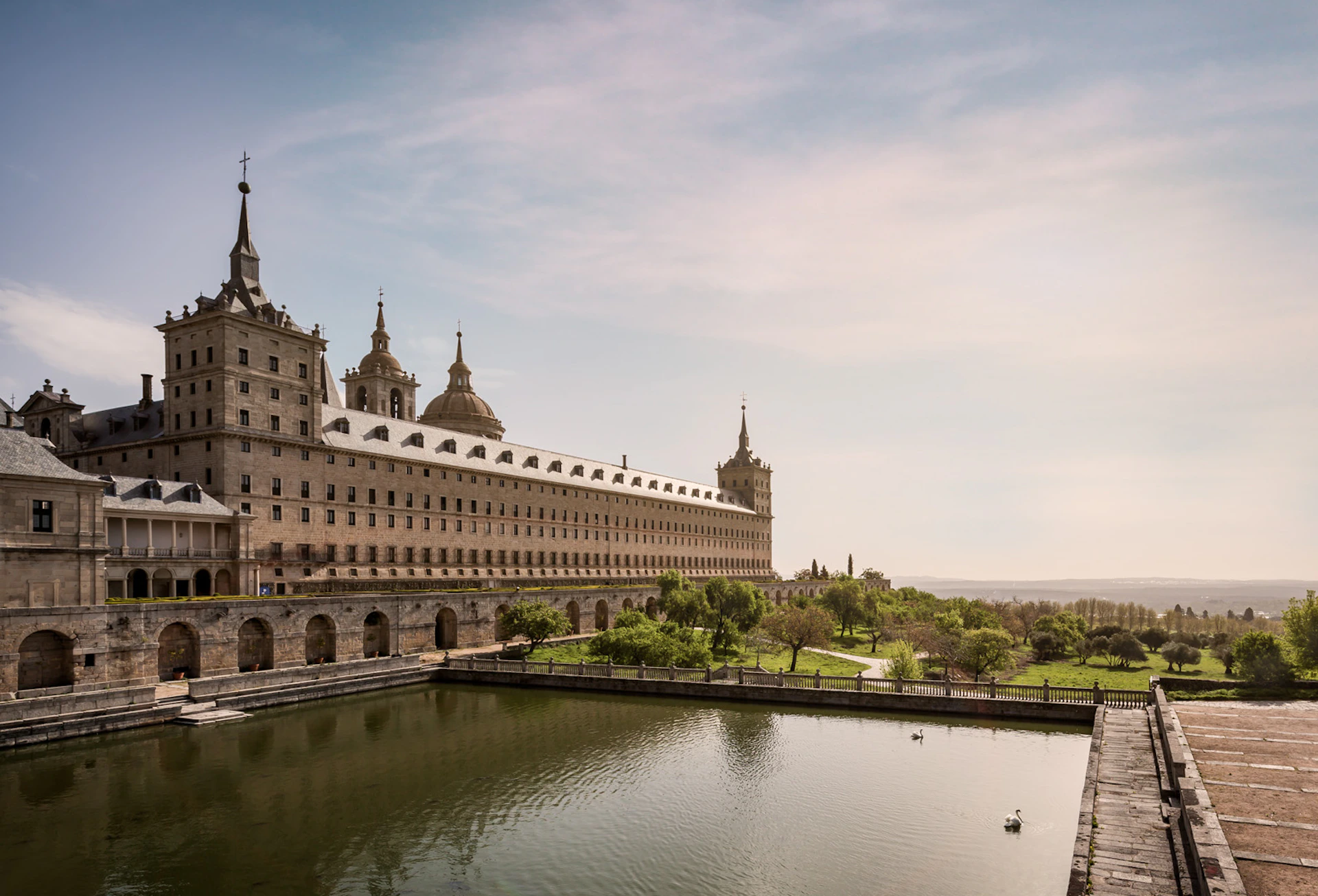 A large historic building with multiple towers is situated beside a rectangular pond bordered by greenery under a partly cloudy sky.
