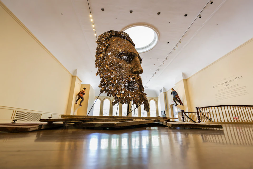 Large metal sculpture of a bearded face with two mannequins posed in dynamic positions behind it, in a well-lit gallery.