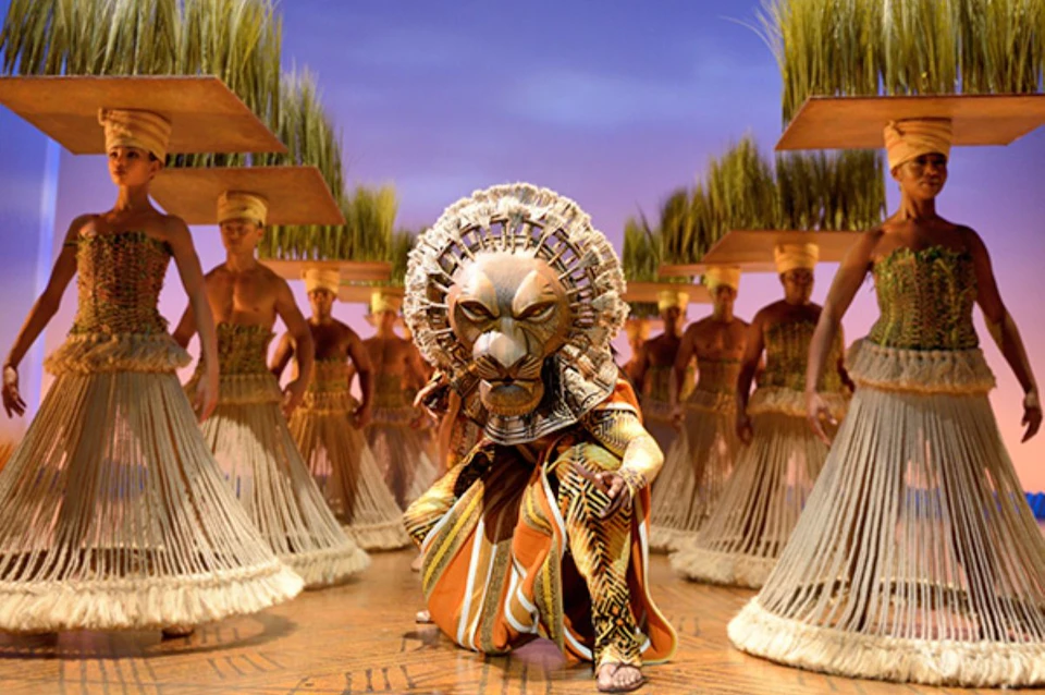 A performer in a lion costume leads a group of dancers in an elaborate stage setting with tall grass props.