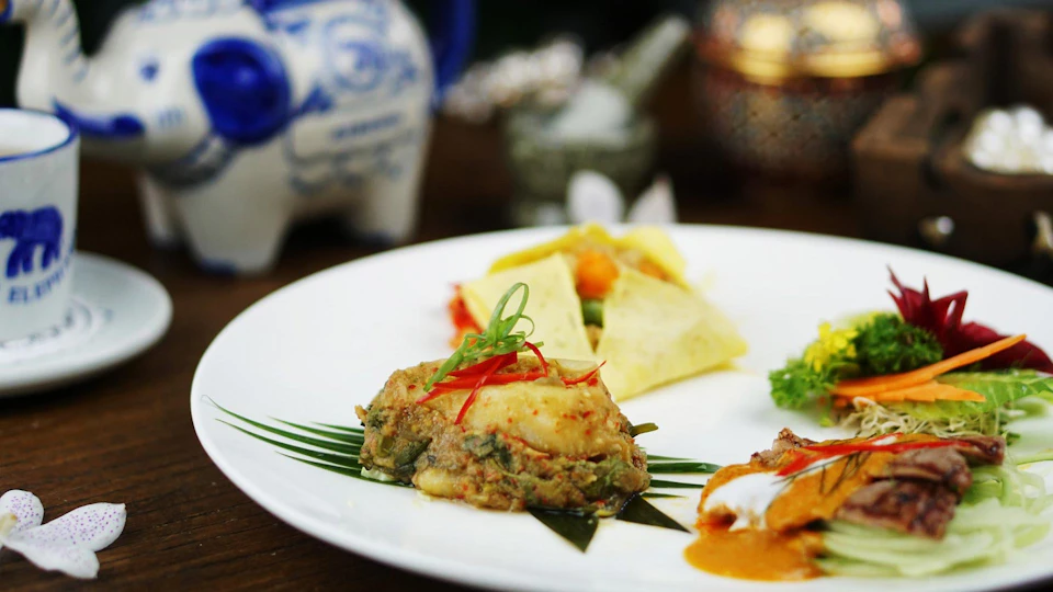A plate with various Thai dishes including a steamed fish cake, garnished with sliced chili and green onions, with decorative elements in the background.
