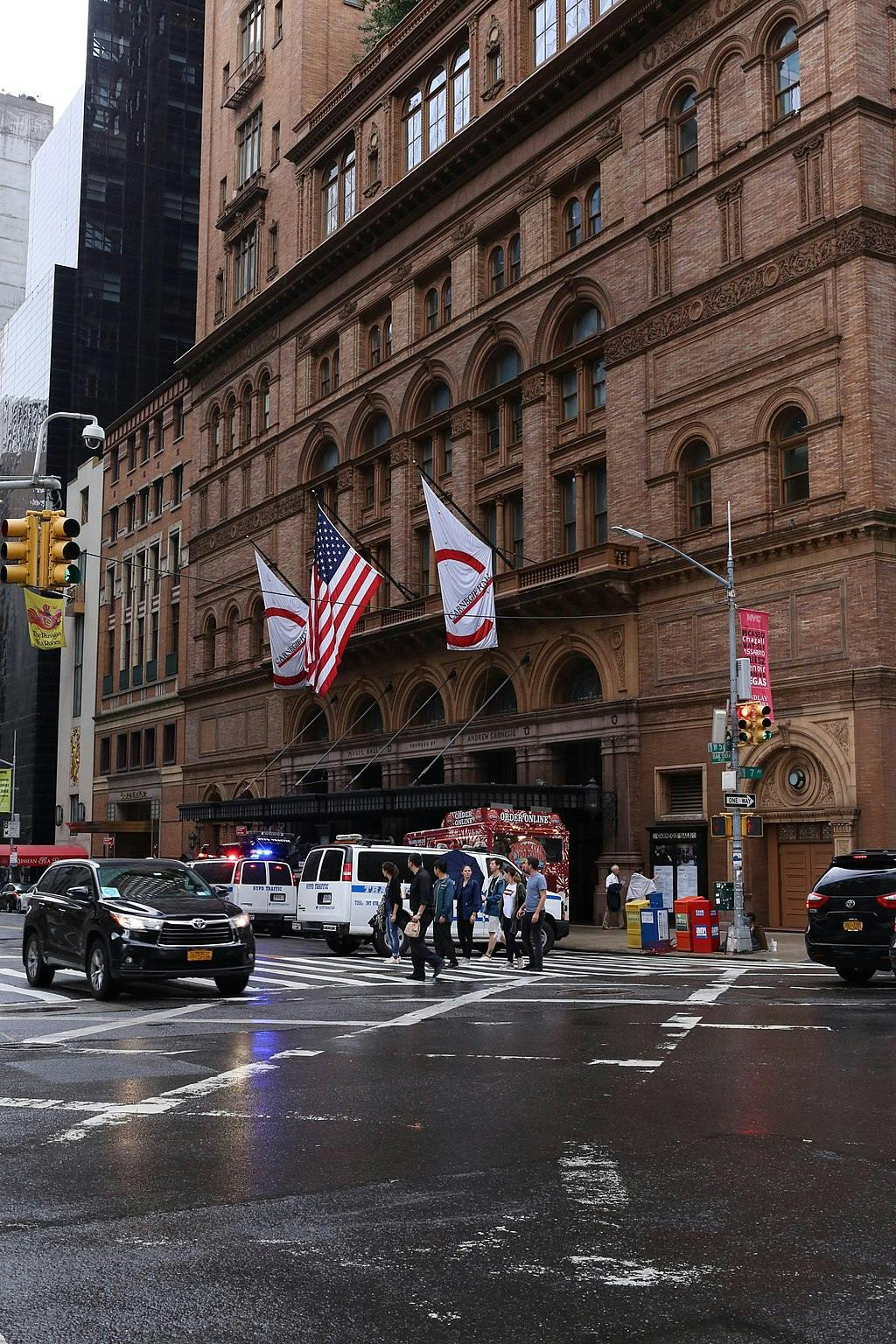 People crossing a street in front of a historic building with American flags, a food truck, cars, and police vans in view.