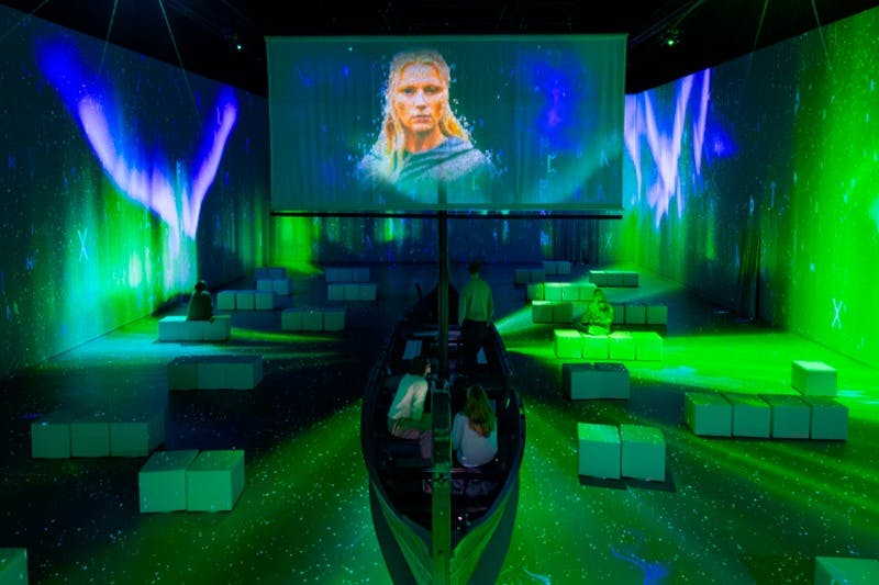 Utställning inomhus med grön belysning, en projektionsskärm som visar en person och en liten båt omgiven av sittkuber.
