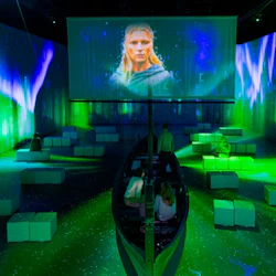 Utställning inomhus med grön belysning, en projektionsskärm som visar en person och en liten båt omgiven av sittkuber.