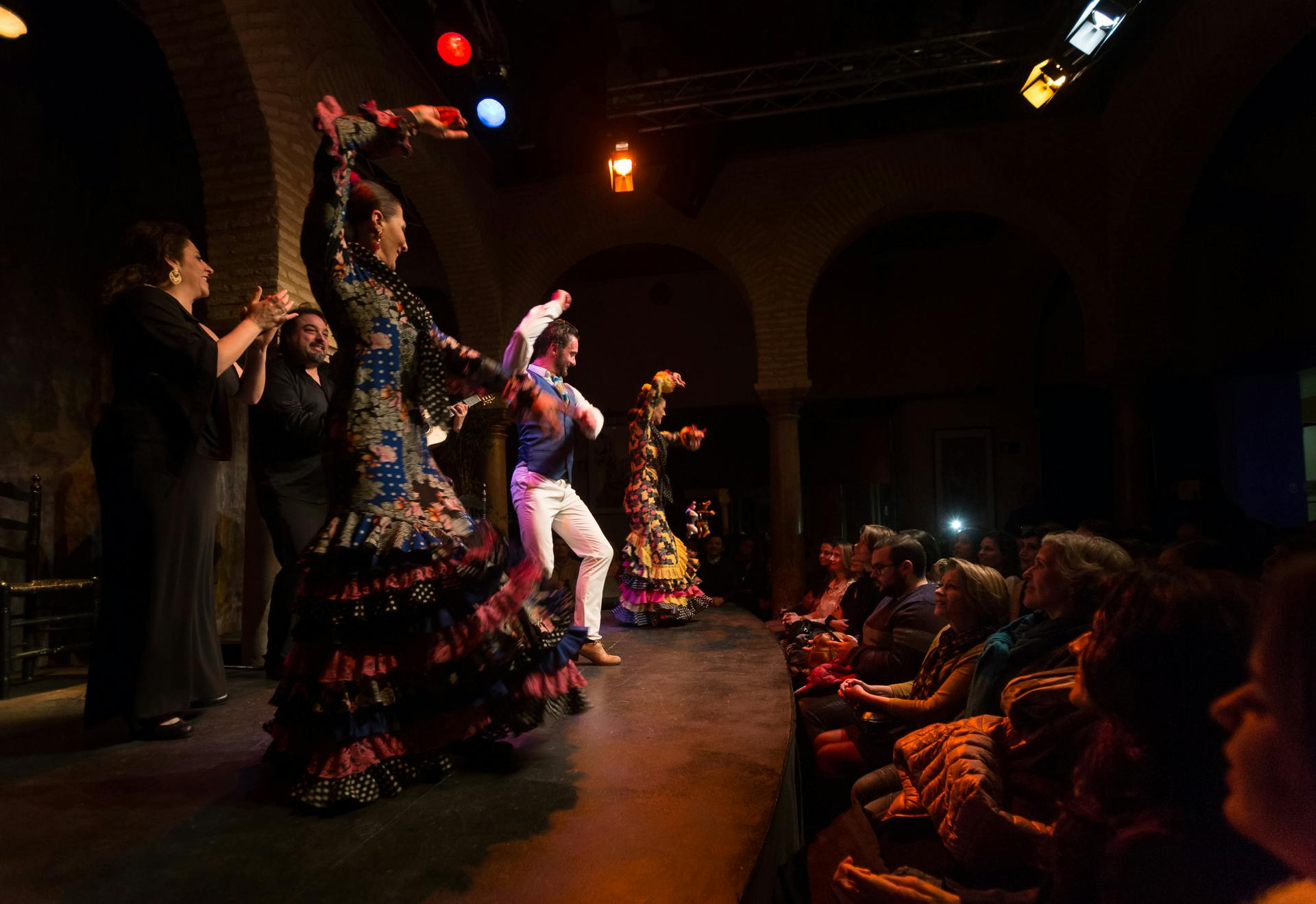 Flamencodansers in kleurrijke jurken treden energiek op het podium op, terwijl een publiek aandachtig toekijkt in een schemerig verlicht theater.