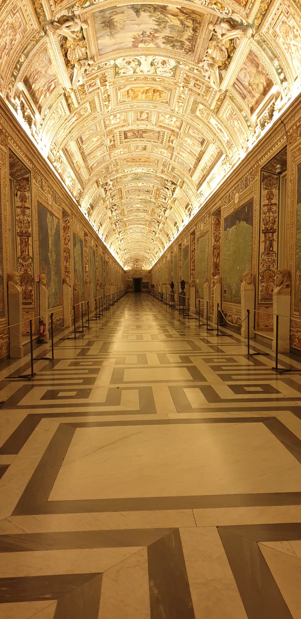 Ornate corridor with illuminated vaulted ceilings, detailed wall murals, patterned marble floor, and sculptures along each side.