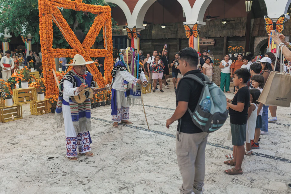 A performer in traditional attire interacts with a crowd, including children, in a plaza decorated with marigolds and butterfly motifs.