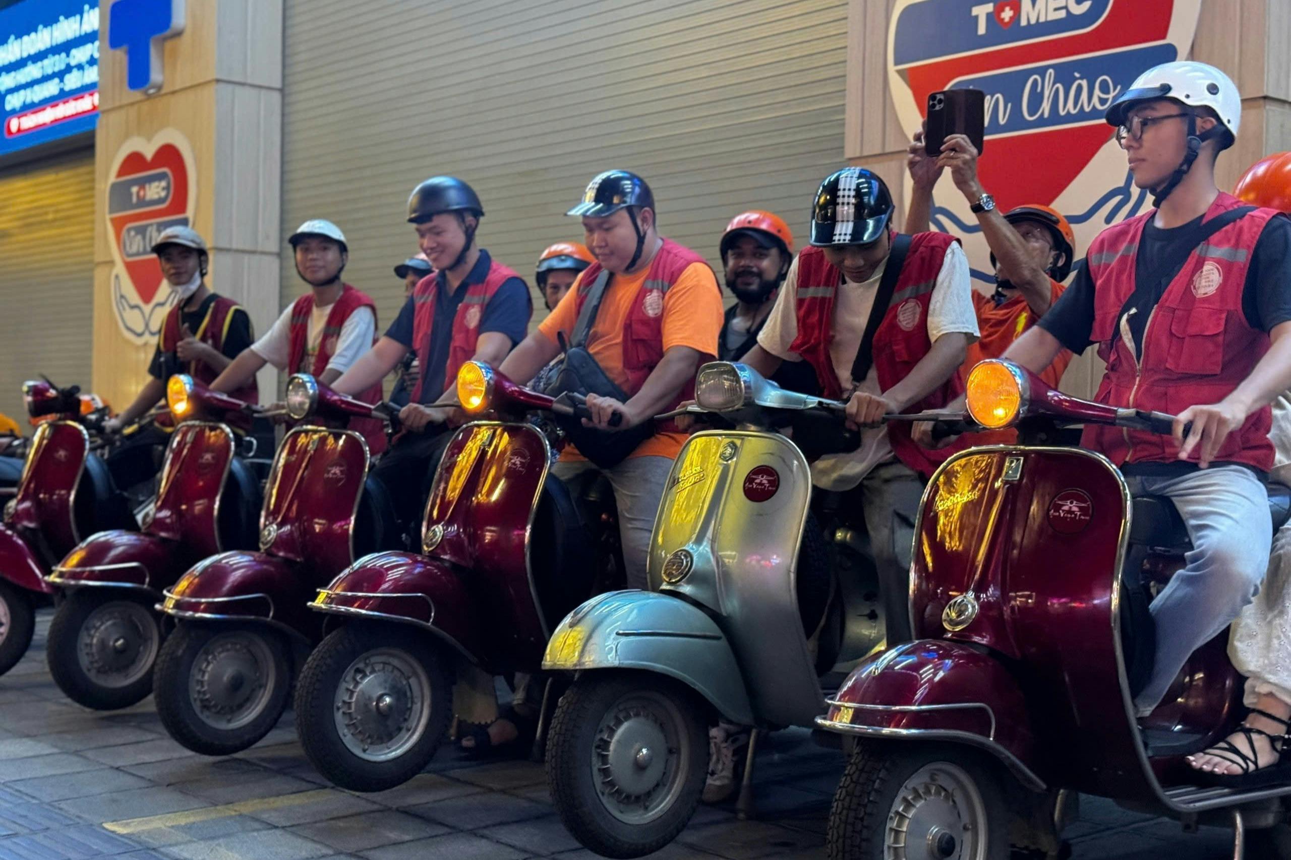 Seven people wearing helmets and orange vests sit on scooters parked in a row on a sidewalk in front of a closed shop.