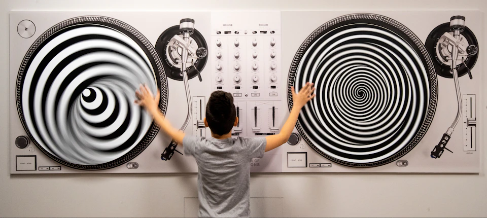 A boy with outstretched arms touches two large, black-and-white, spiral-patterned turntable illustrations on a wall.