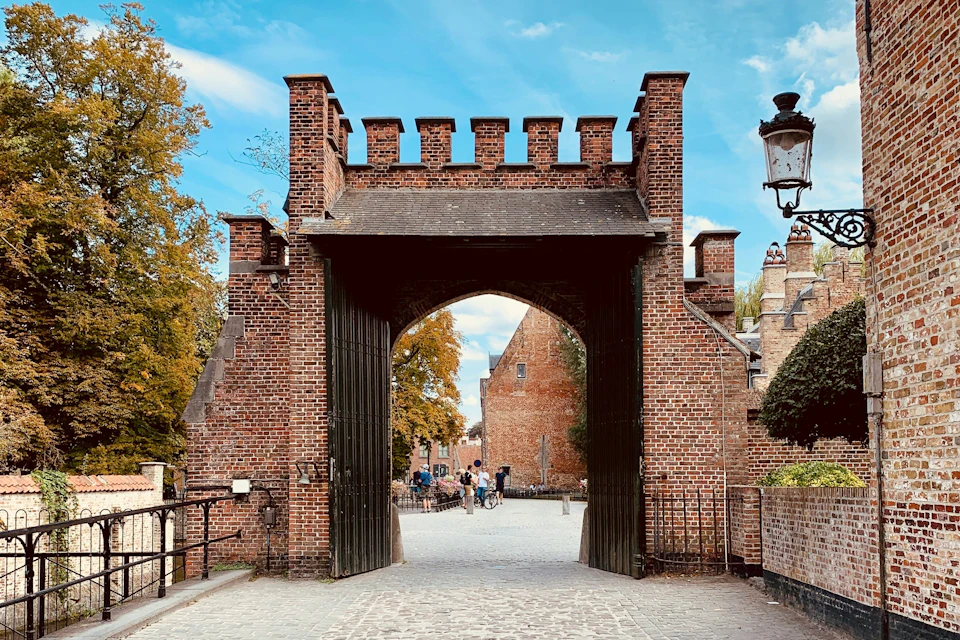 An arched brick gateway leads to a cobblestone street with people in the background, surrounded by autumn trees and old buildings.