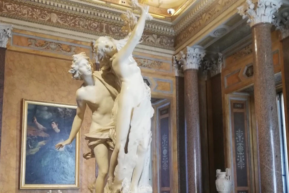 A marble sculpture of two figures in a dramatic pose, displayed in an ornate room featuring classical architectural details.