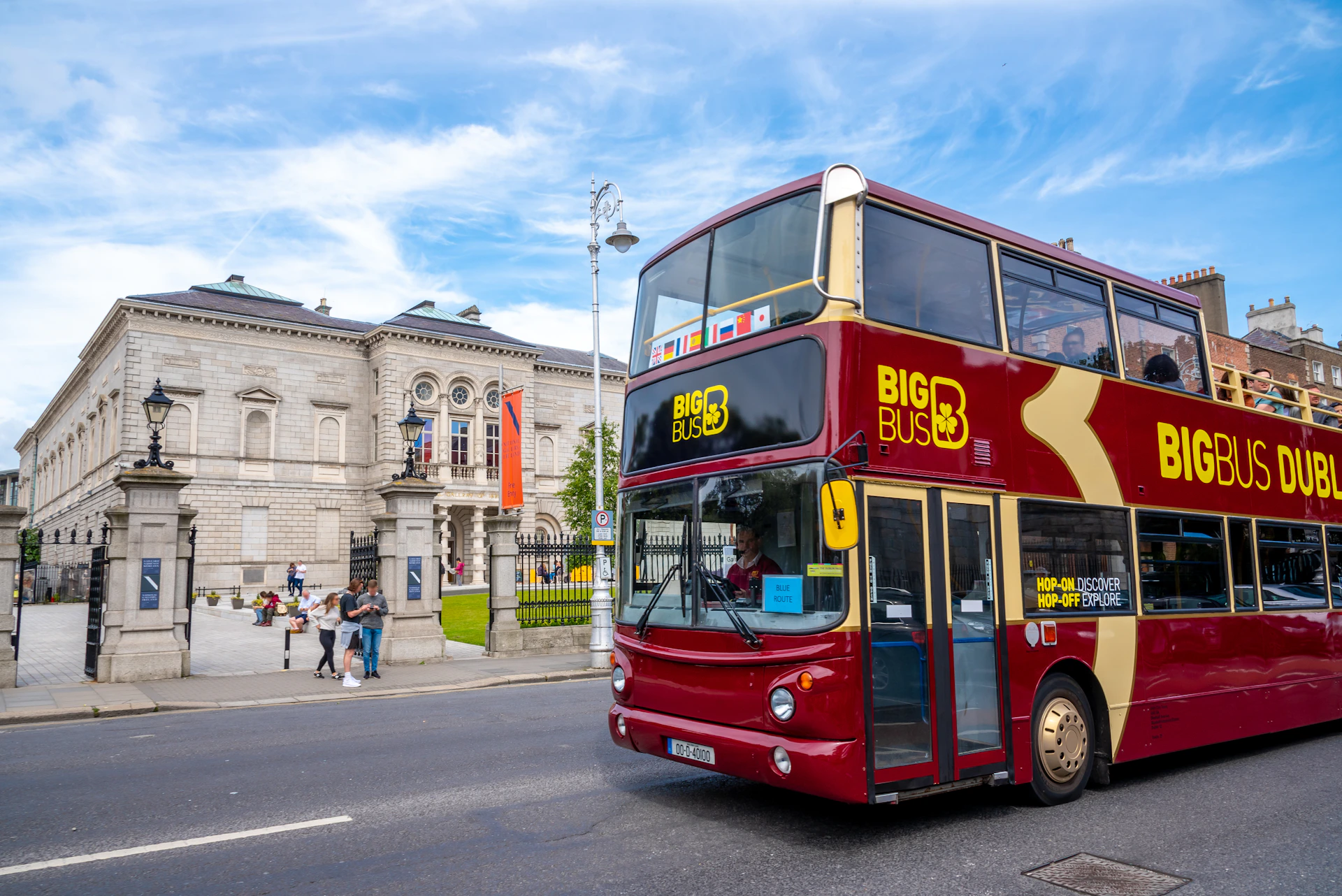A red double-decker "Big Bus Tours" vehicle is parked in front of an ornate building with people nearby and a lamppost visible.